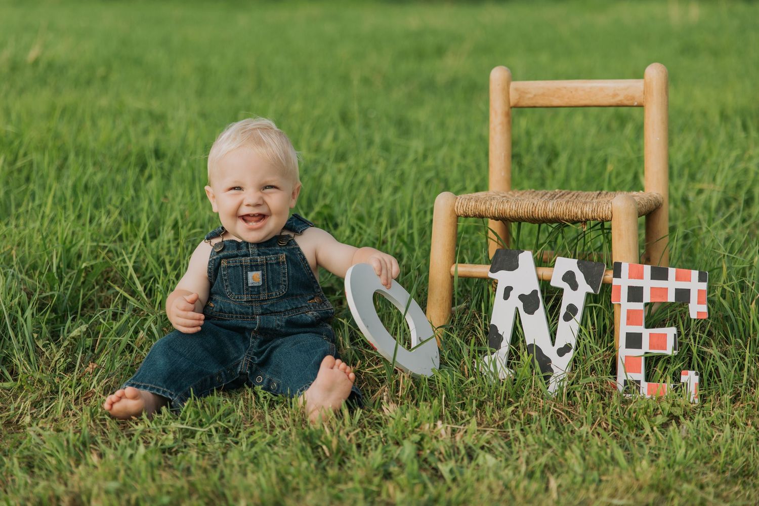 Laughing baby in overalls sits on grass next to wooden chair and decorative blocks.