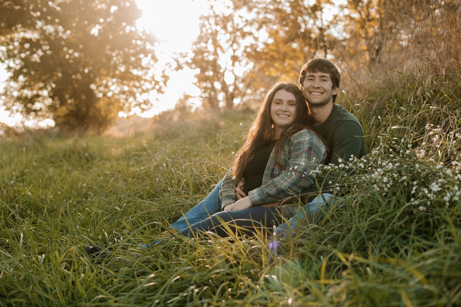 Two people sitting in tall grass on a hillside during golden hour with small white wildflowers in the foreground.
