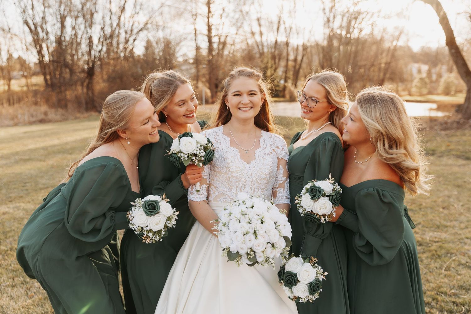 Wedding party wearing emerald green dresses and holding white floral bouquets pose together in a sunlit autumn field.