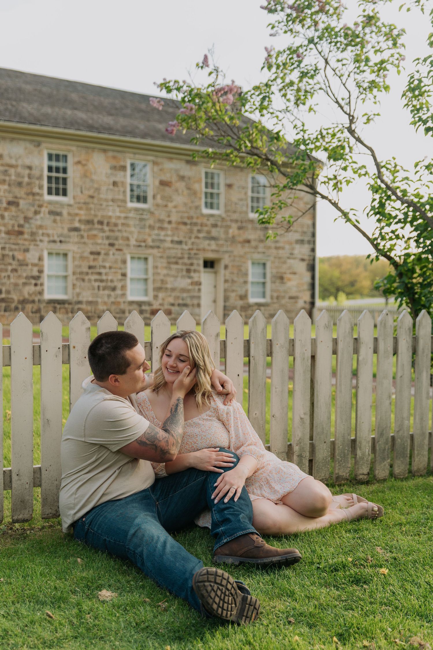 A casual engagement photo of a couple relaxing on grass in front of a historic stone building in Central Pennsylvania
