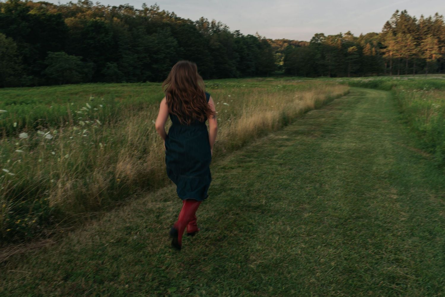 Figure in navy dress and red boots walking along grassy countryside path at sunset.