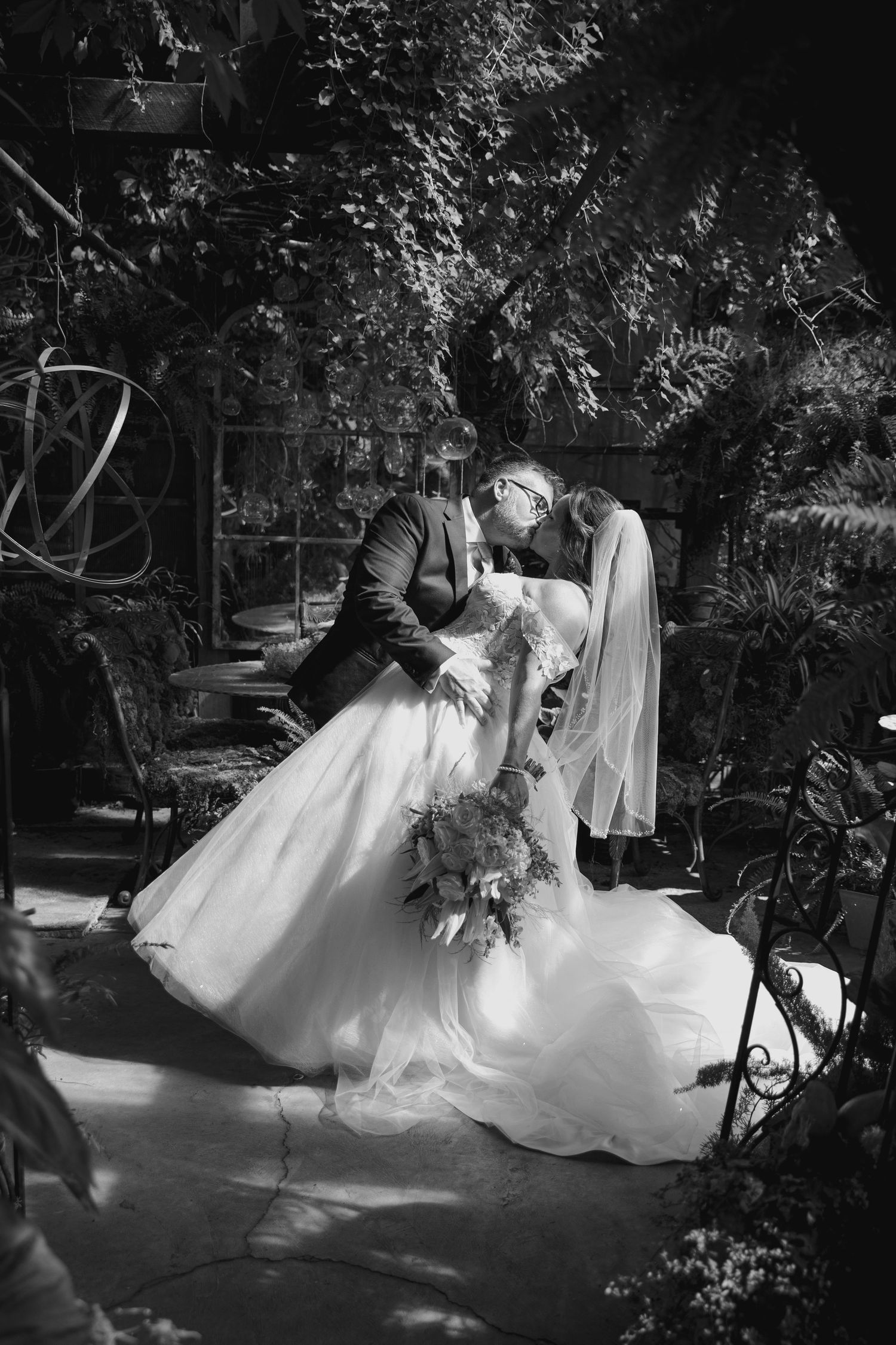 A dramatic black and white wedding photo captured in a lush garden setting with decorative ironwork.