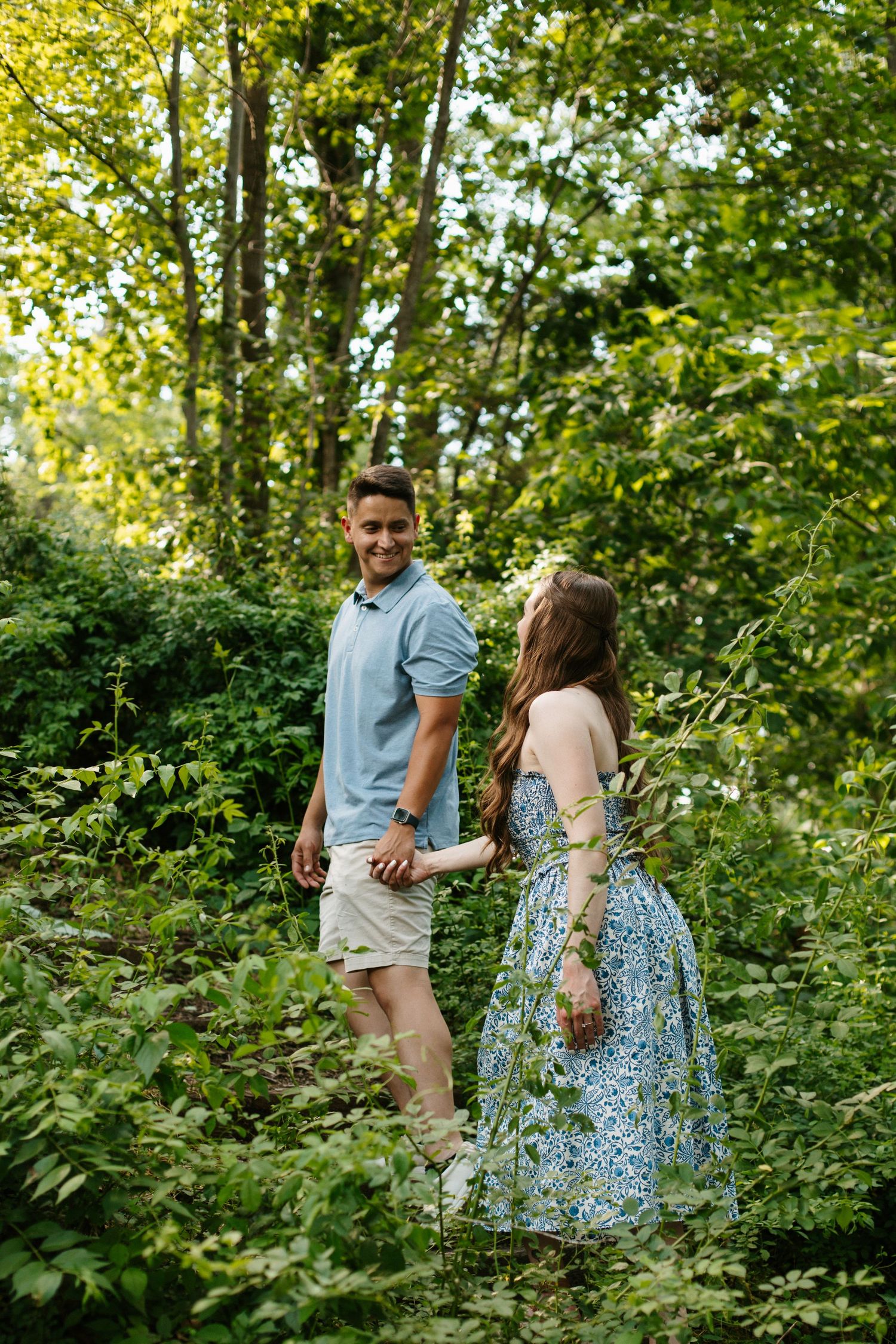 A smiling couple holds hands while walking through a lush green forest, the woman wearing a blue floral dress.