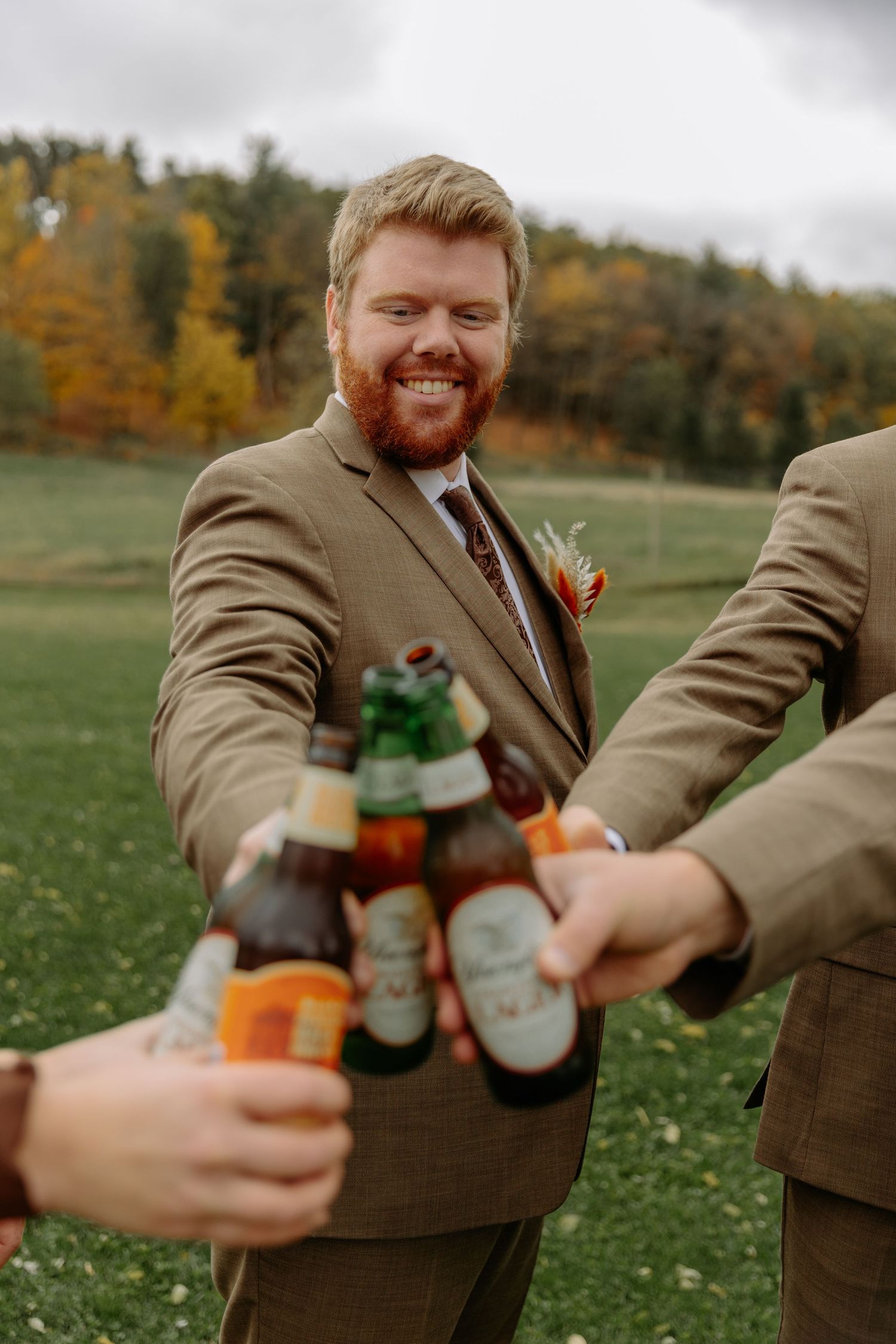 Groomsmen in tan suits toasting and laughing with beer bottles at an outdoor wedding reception.