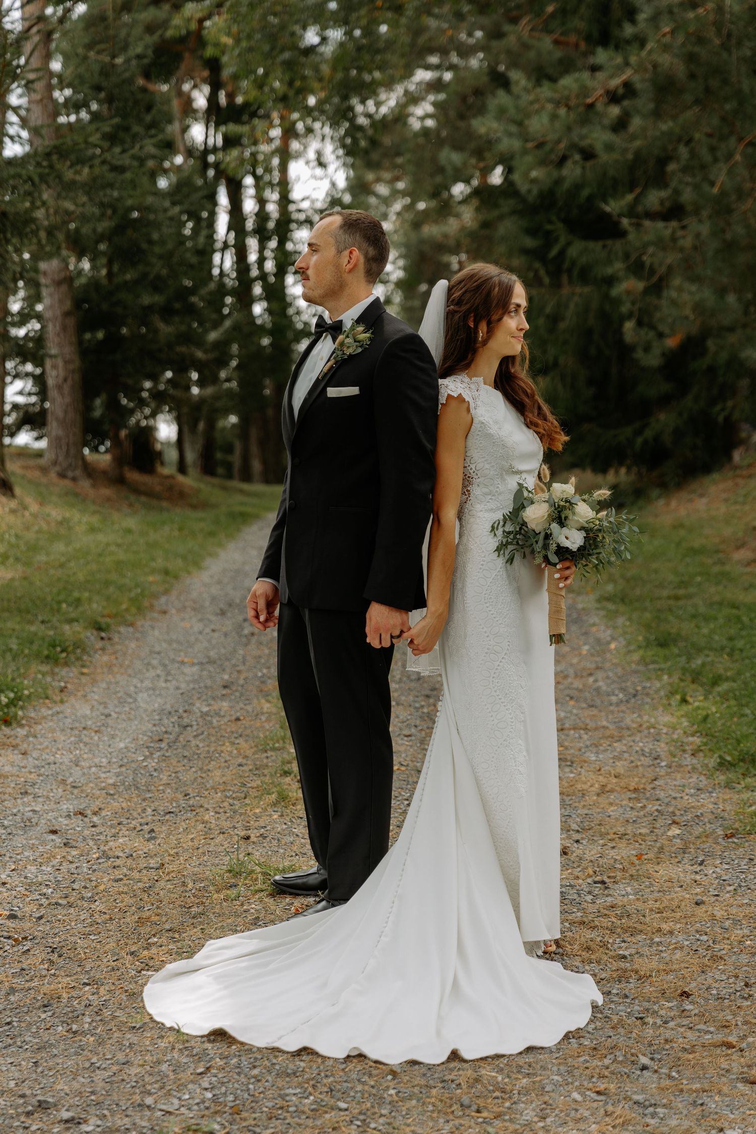 A wedding couple stands back-to-back on a gravel path surrounded by evergreen trees.
