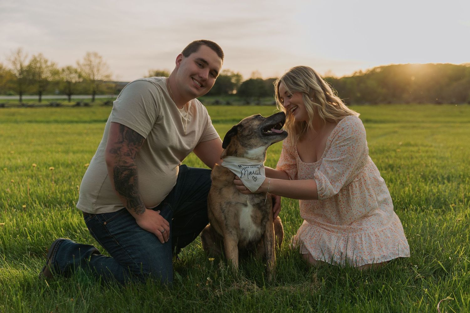 Couple pets their dog during sunset engagement session in grassy field.
