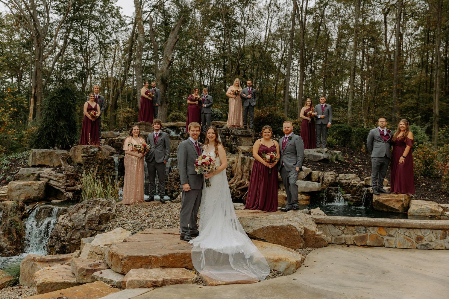 Wedding party poses on rustic stone steps in a wooded outdoor venue with burgundy and neutral colored attire.