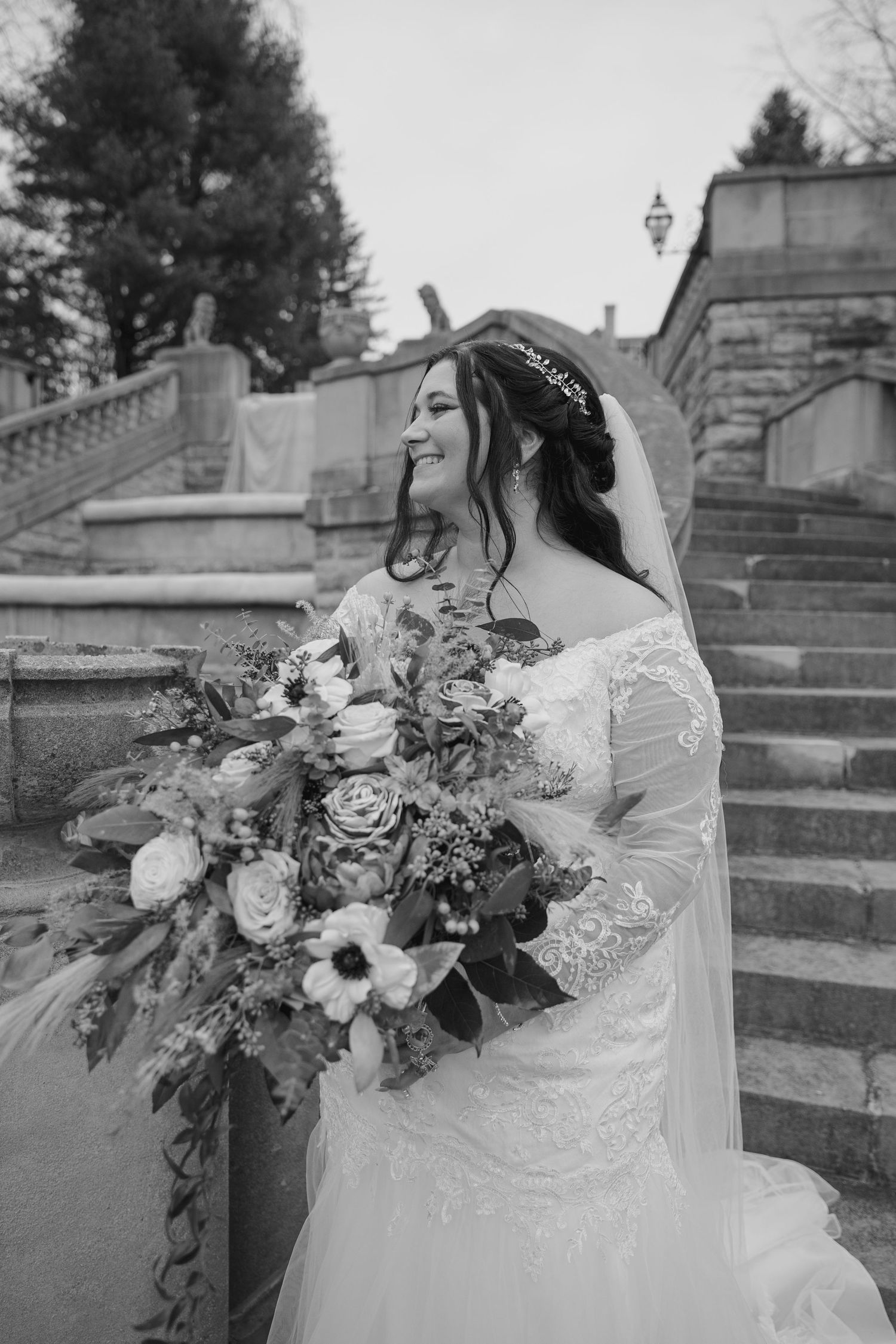 Bride in lace gown holds luxurious floral bouquet on stone steps in black and white photo.