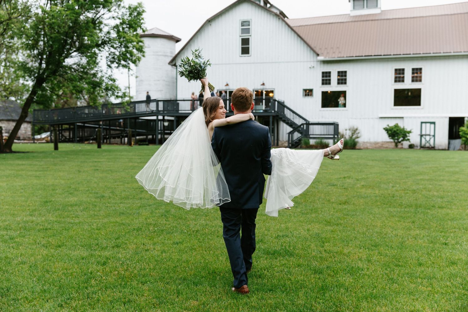 Groom carrying bride in white wedding dress across green lawn toward white barn venue.