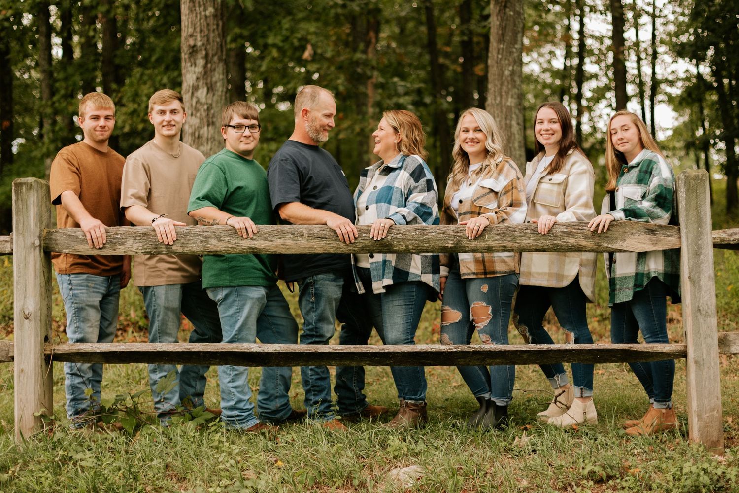 A group of people in casual attire leaning against a wooden fence in a rustic outdoor setting with trees in background.