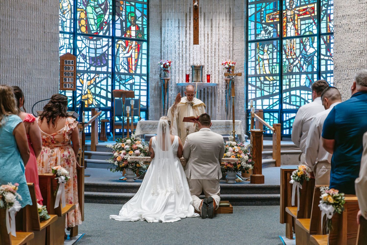 Bride and groom kneeling at church altar during wedding ceremony with stained glass windows and guests standing nearby.
