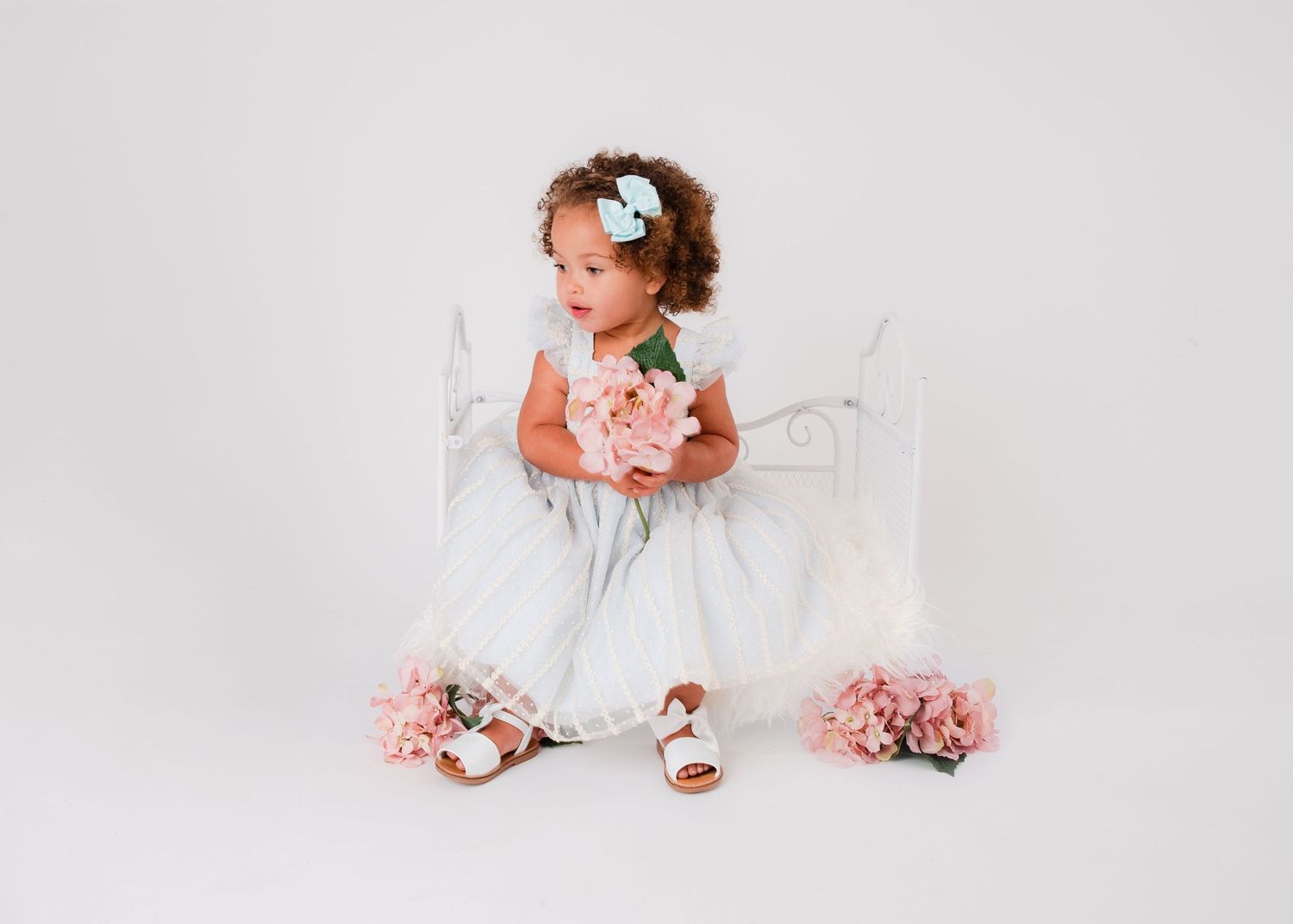 A toddler in white dress sits among scattered pink roses on white background.