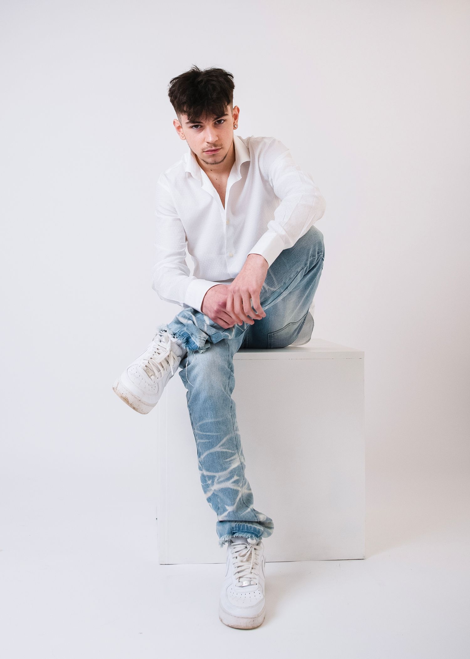 Young model in white shirt and distressed jeans poses on white cube in studio setting.