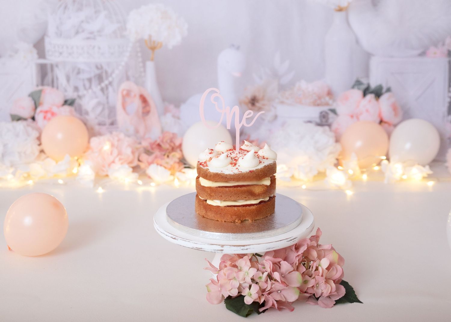 Pink and white party decorations with string lights and cake display on white backdrop.