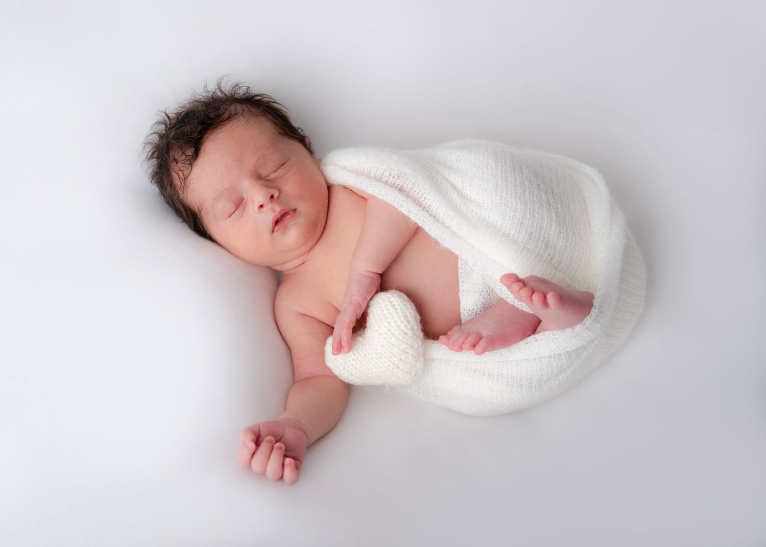 Newborn baby wrapped in soft white swaddle against white backdrop.