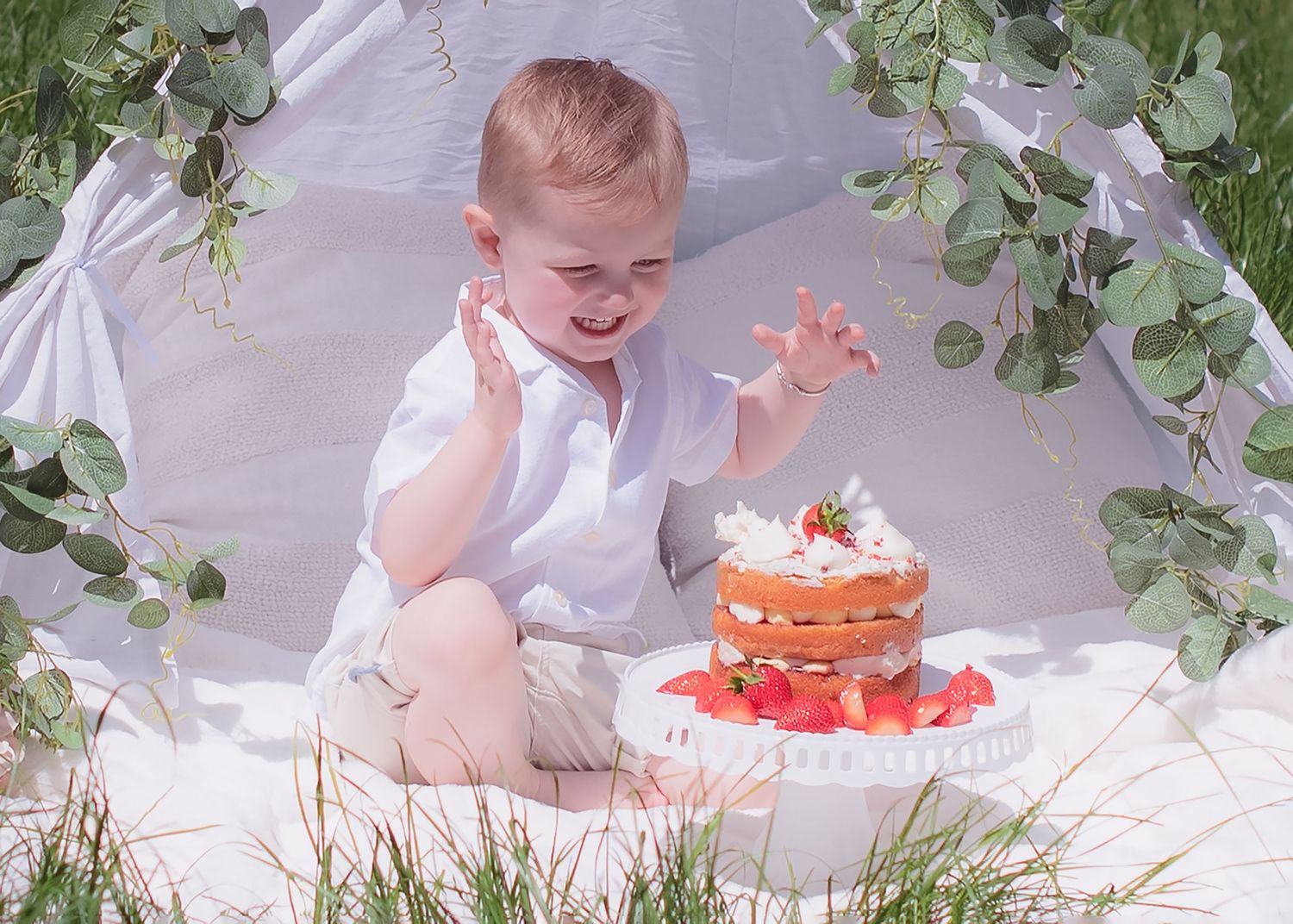 Baby celebrating first birthday with a decorated cake and greenery backdrop.