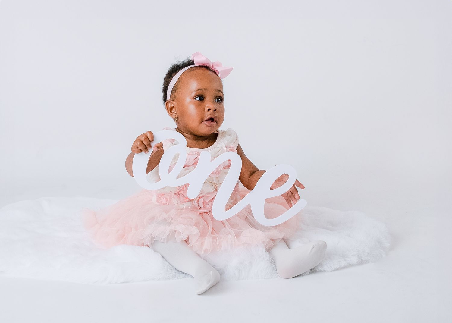 Newborn baby wearing white outfit and headband lying on soft white backdrop in ethereal studio lighting.