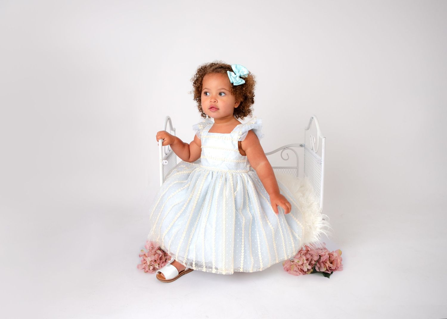 A small child in a white dress poses with pink flowers against a bright white background.