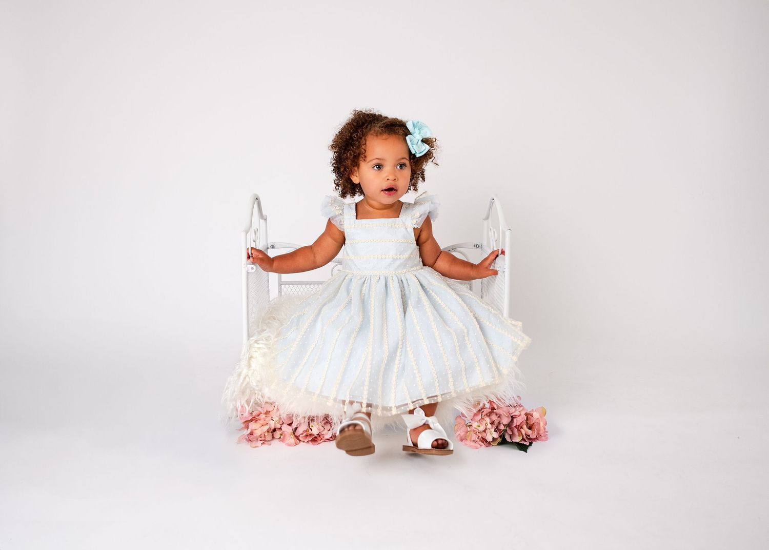Child in white dress poses with pink flowers in bright studio setting.