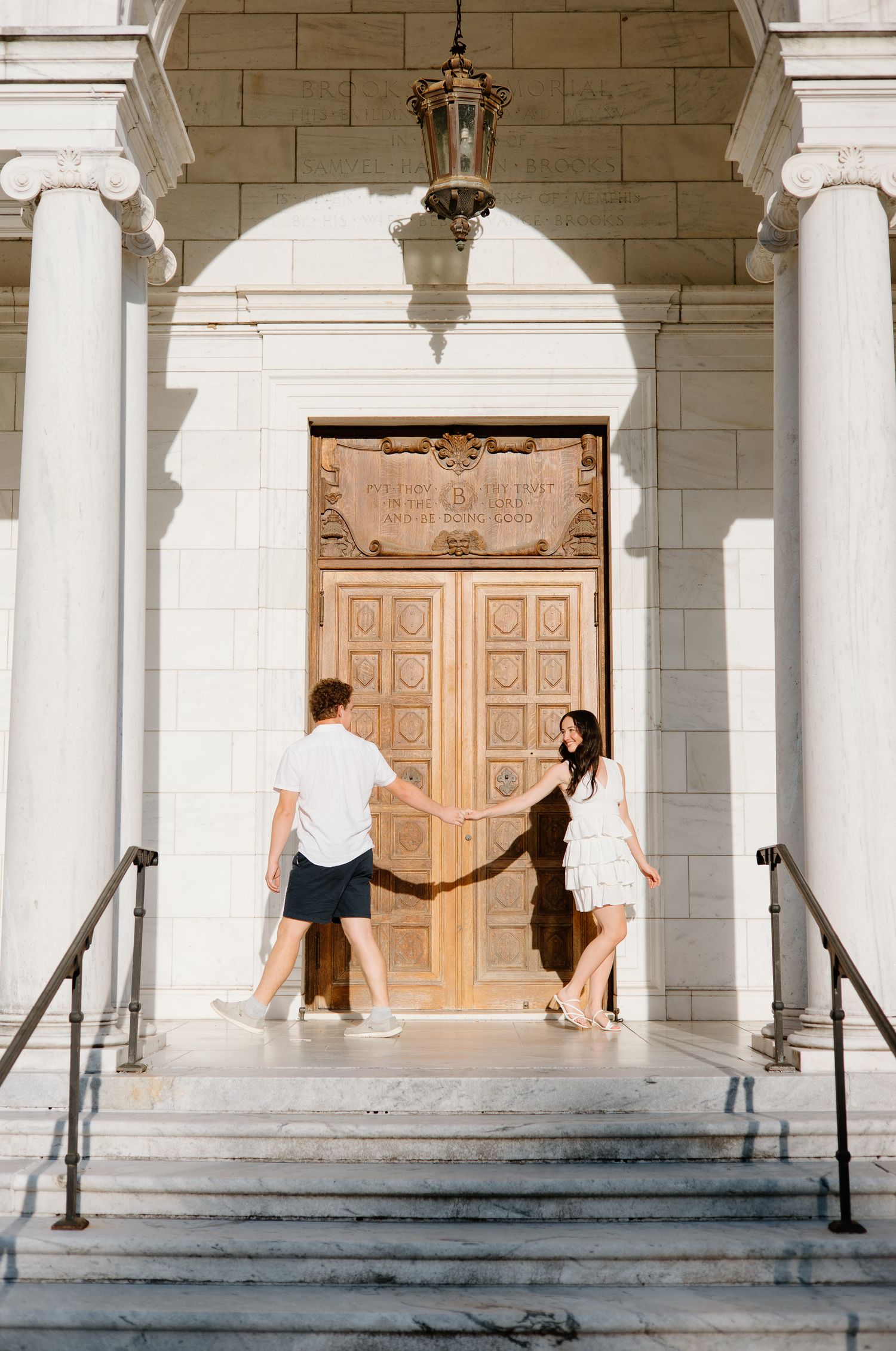 Playful couple posing at grand entrance with white columns and ornate wooden door.