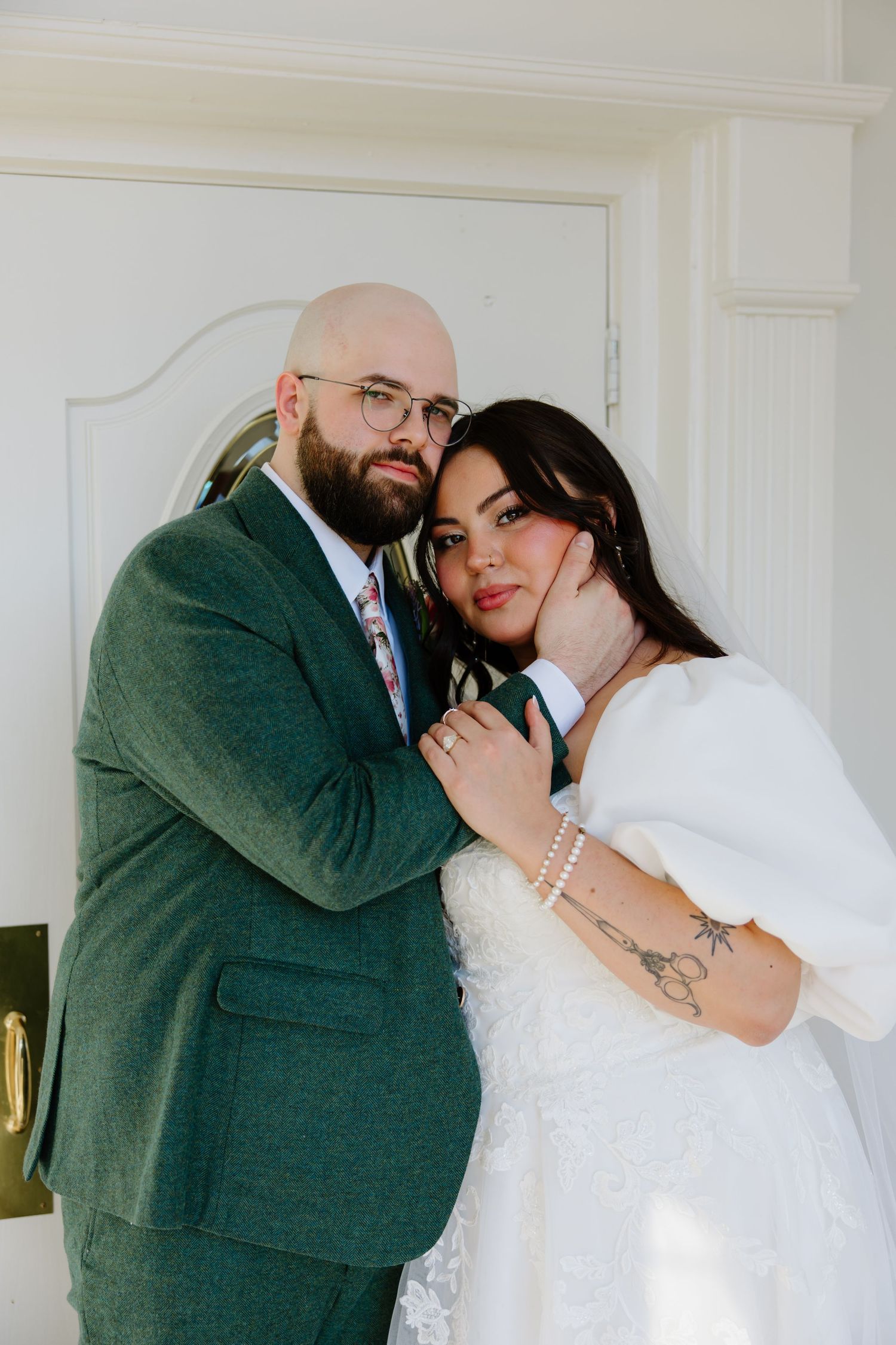 Romantic indoor wedding portrait of couple in white dress and green suit against white wall.