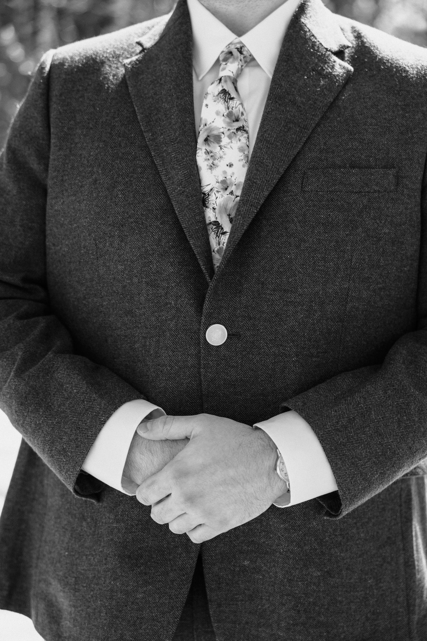 Black and white photo showing torso view of suit jacket with floral tie and clasped hands in formal attire. Wedding Photographer