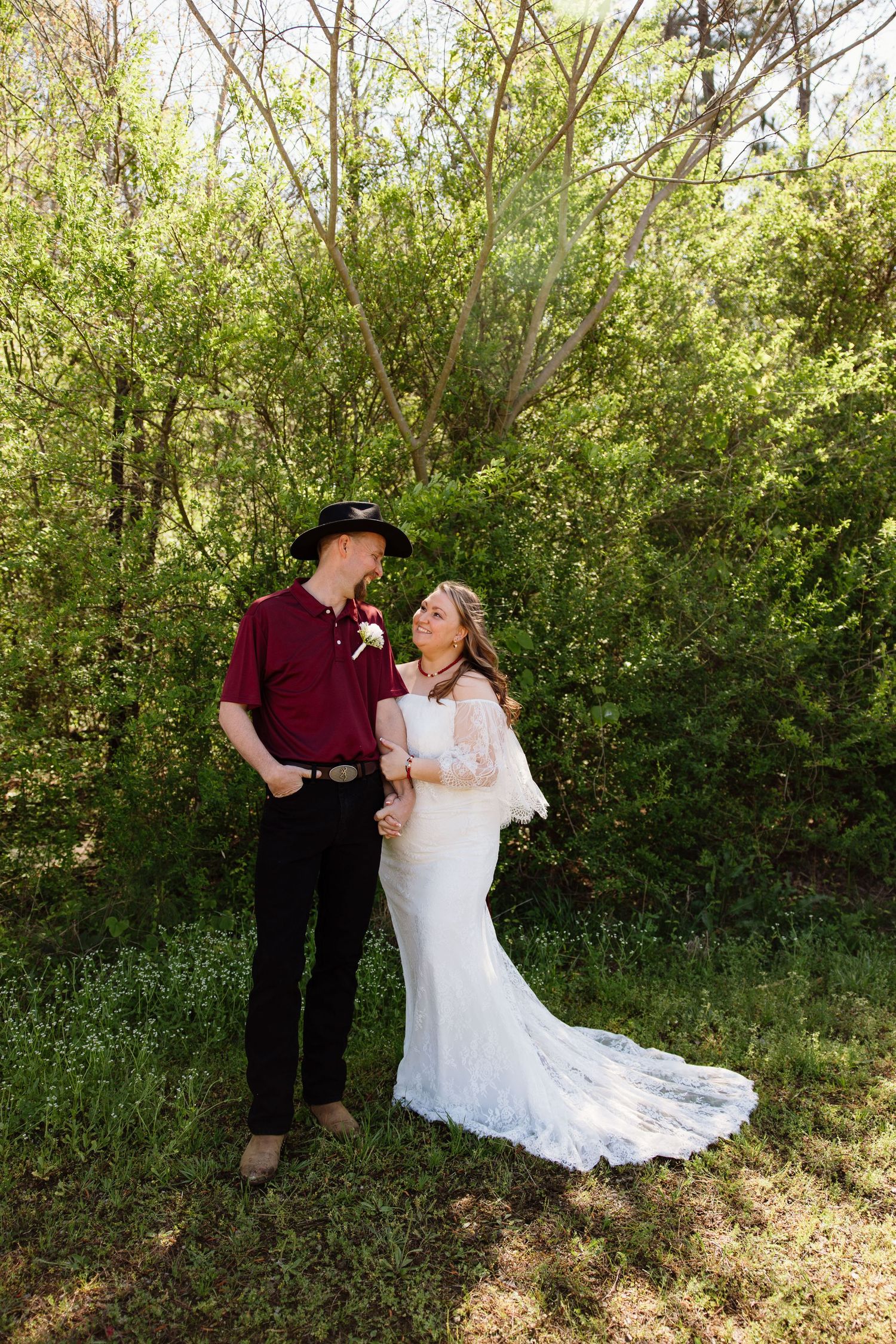A couple in wedding attire poses outdoors, with the groom in burgundy and bride in lace.