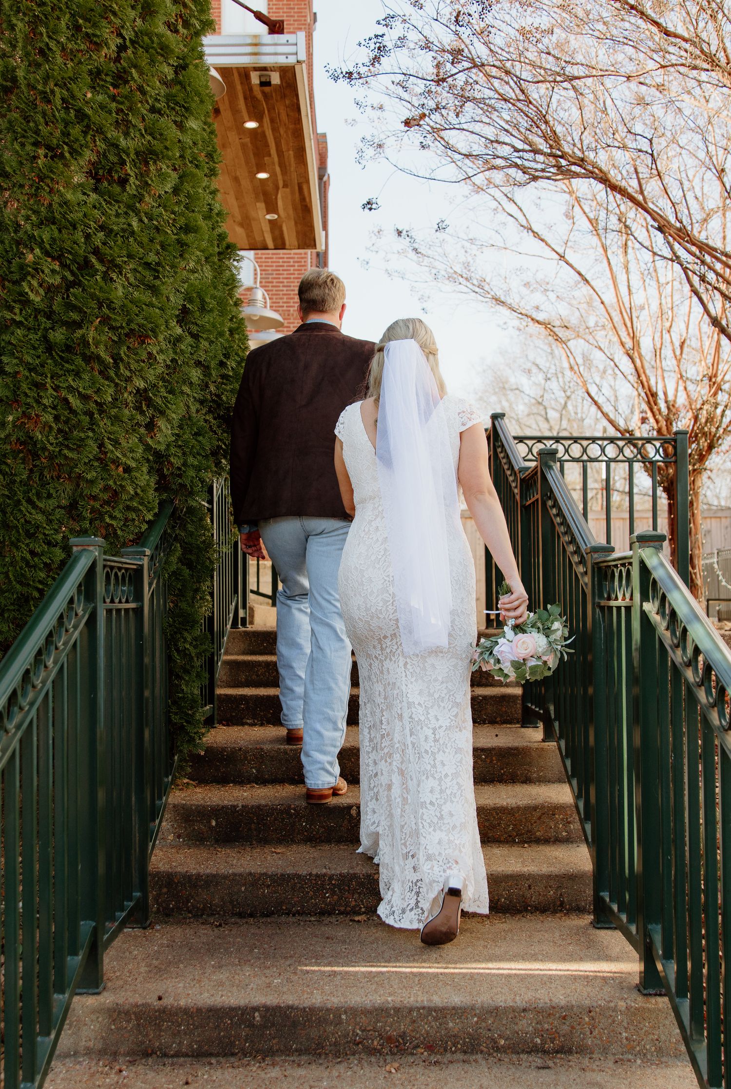 A couple climbs stone steps with green railings while wearing wedding attire on a sunny autumn day.