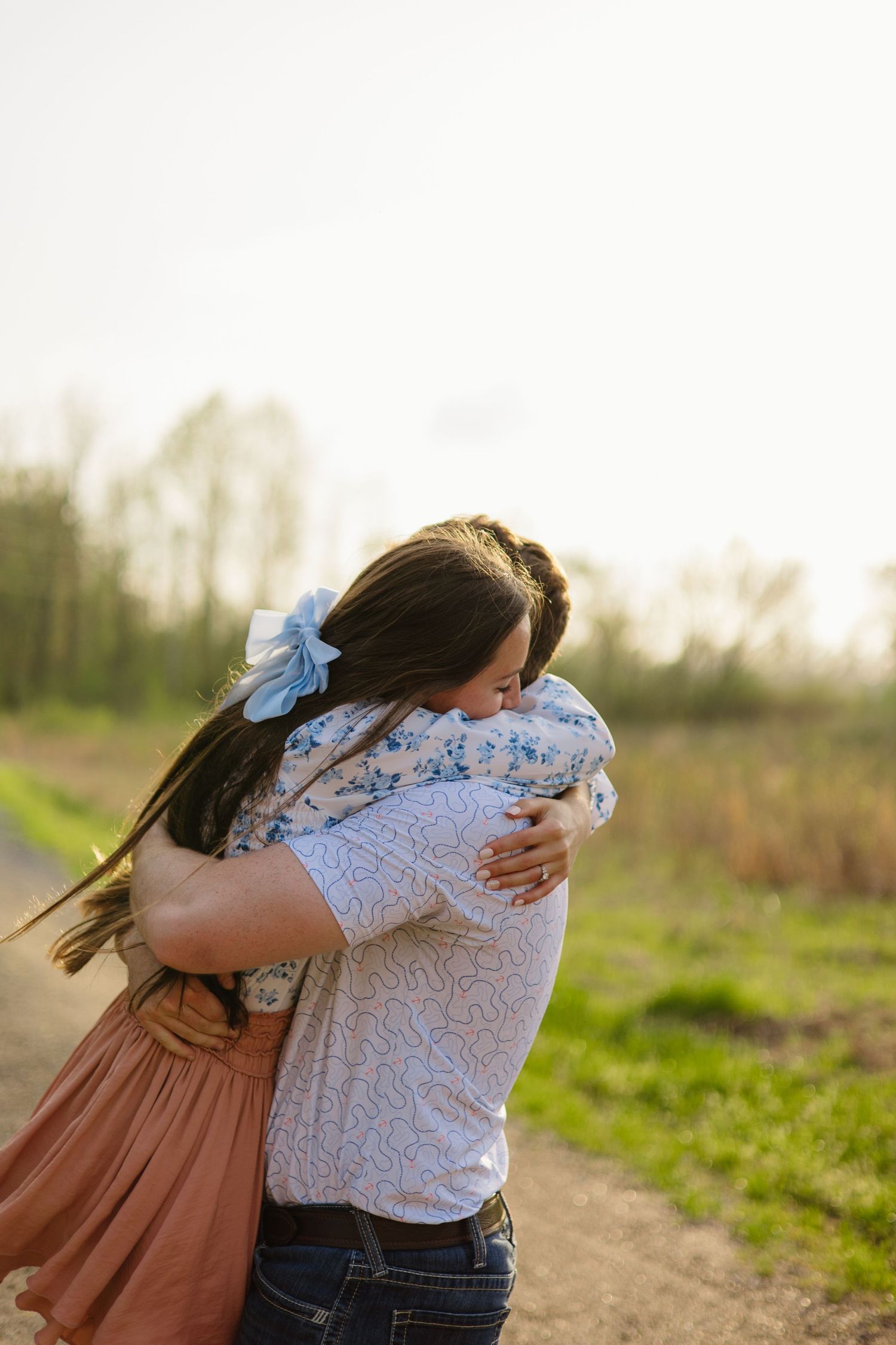 Two people embrace in a tender hug outdoors with sunlit grass in the background.