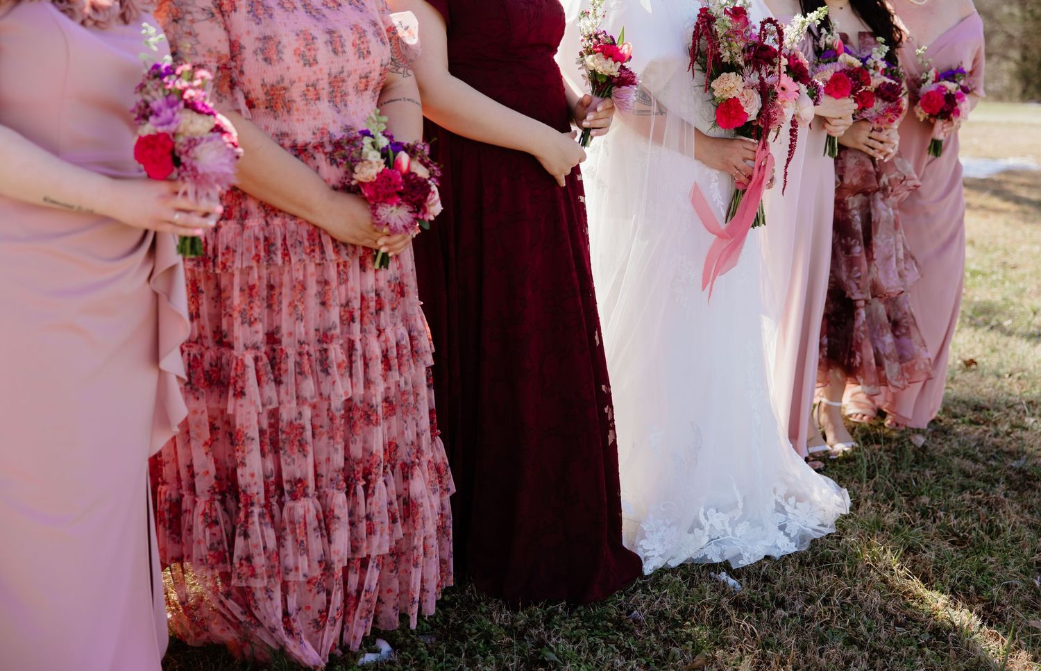 Wedding party holding pink and burgundy bouquets in a row against natural outdoor lighting.