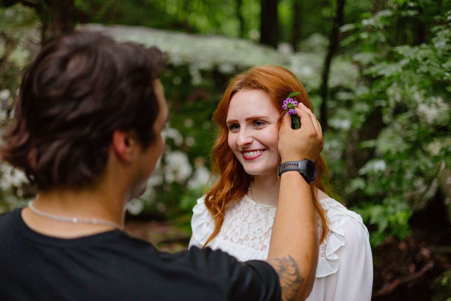 A person adjusts another's hair in a serene outdoor setting with green foliage in the background.