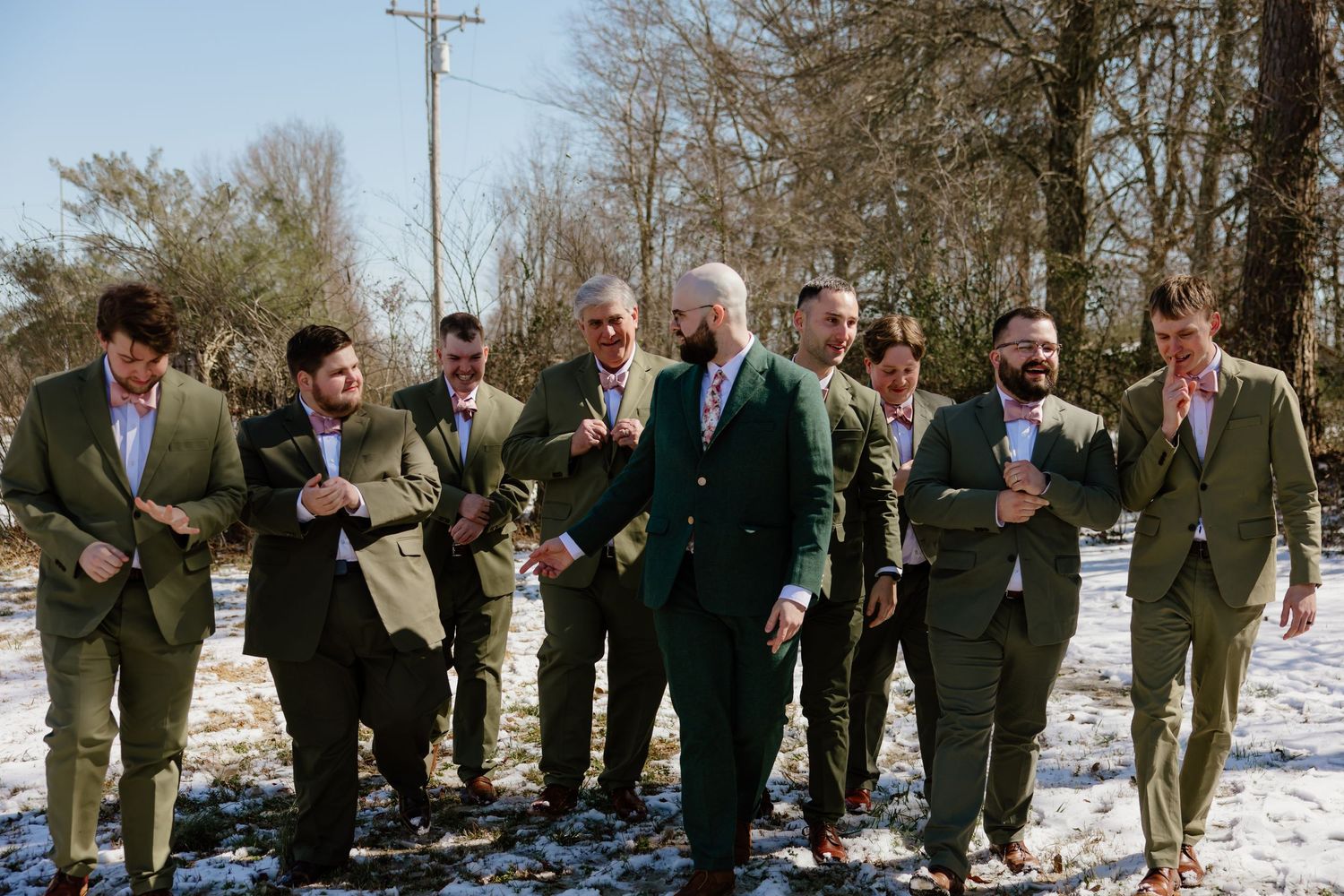 A wedding party wearing green and olive suits stands together on snowy ground in winter sunlight.