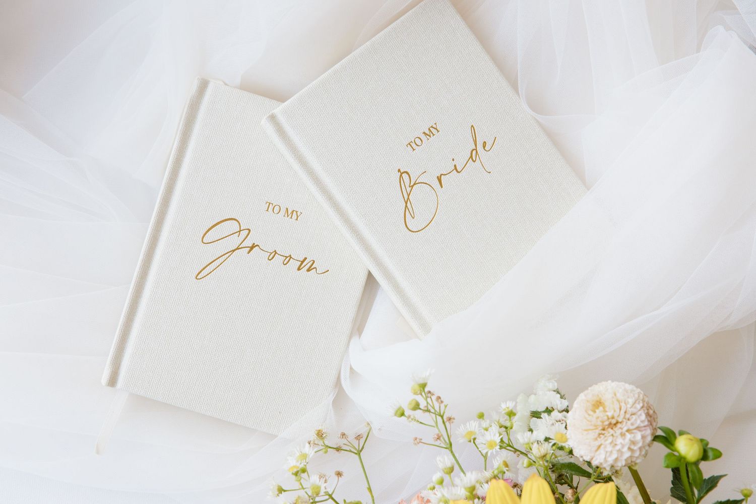 White wedding vow books with gold lettering saying Groom and Bride beside soft flowers.
