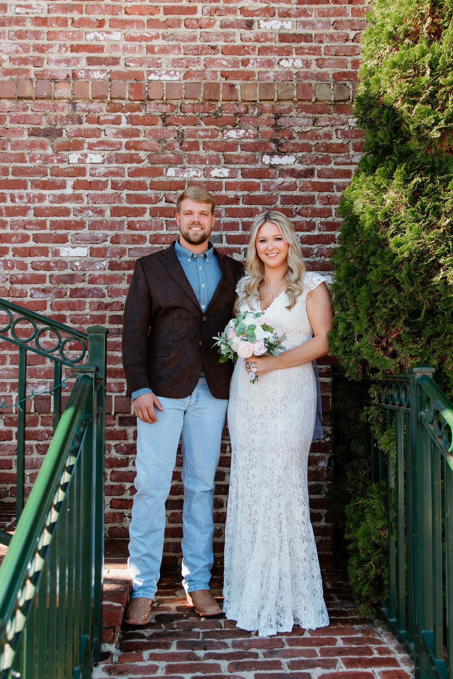 A couple poses together on brick steps with greenery and a rustic brick wall backdrop on their wedding day.