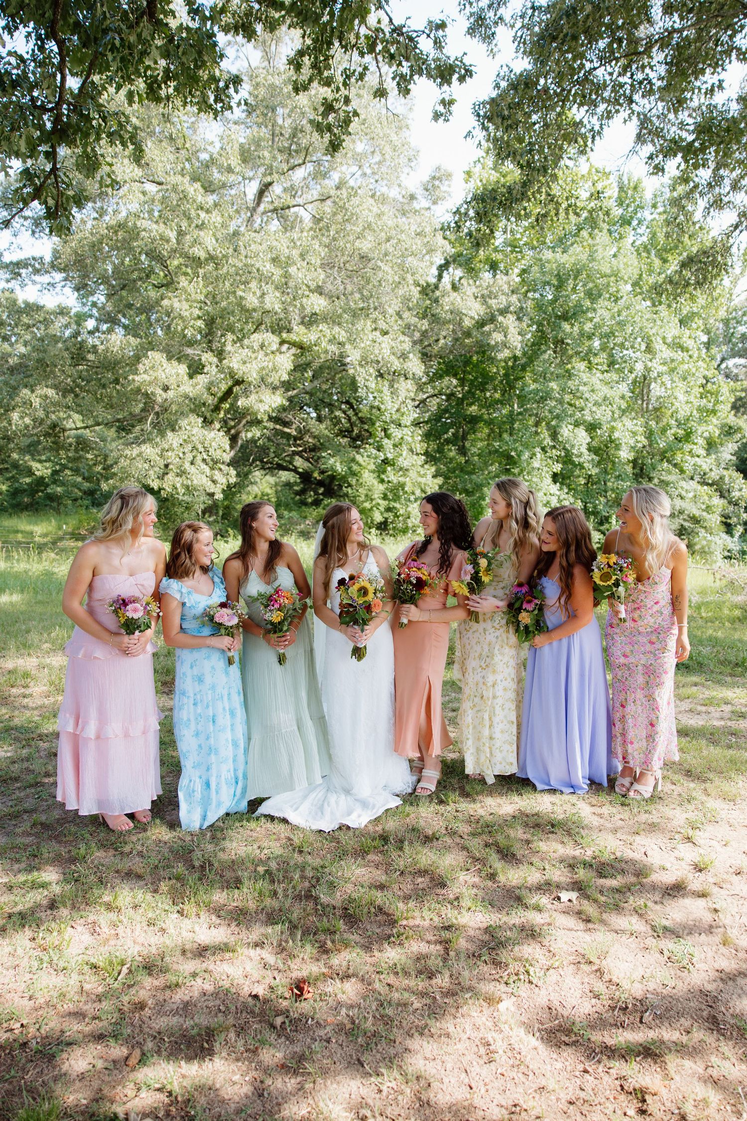 Bridesmaids in pastel dresses stand together holding sunflower bouquets outdoors.