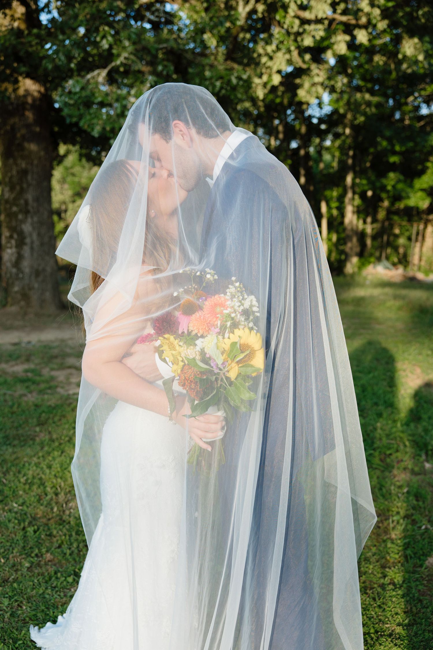 A couple shares a romantic kiss under a flowing veil while holding a vibrant sunflower bouquet in an outdoor garden setting.