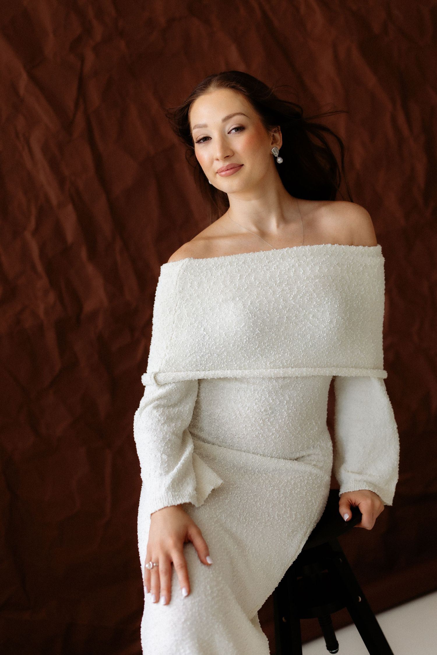 Elegant off-shoulder white knit dress with textured fabric against dark brown backdrop in studio portrait.
