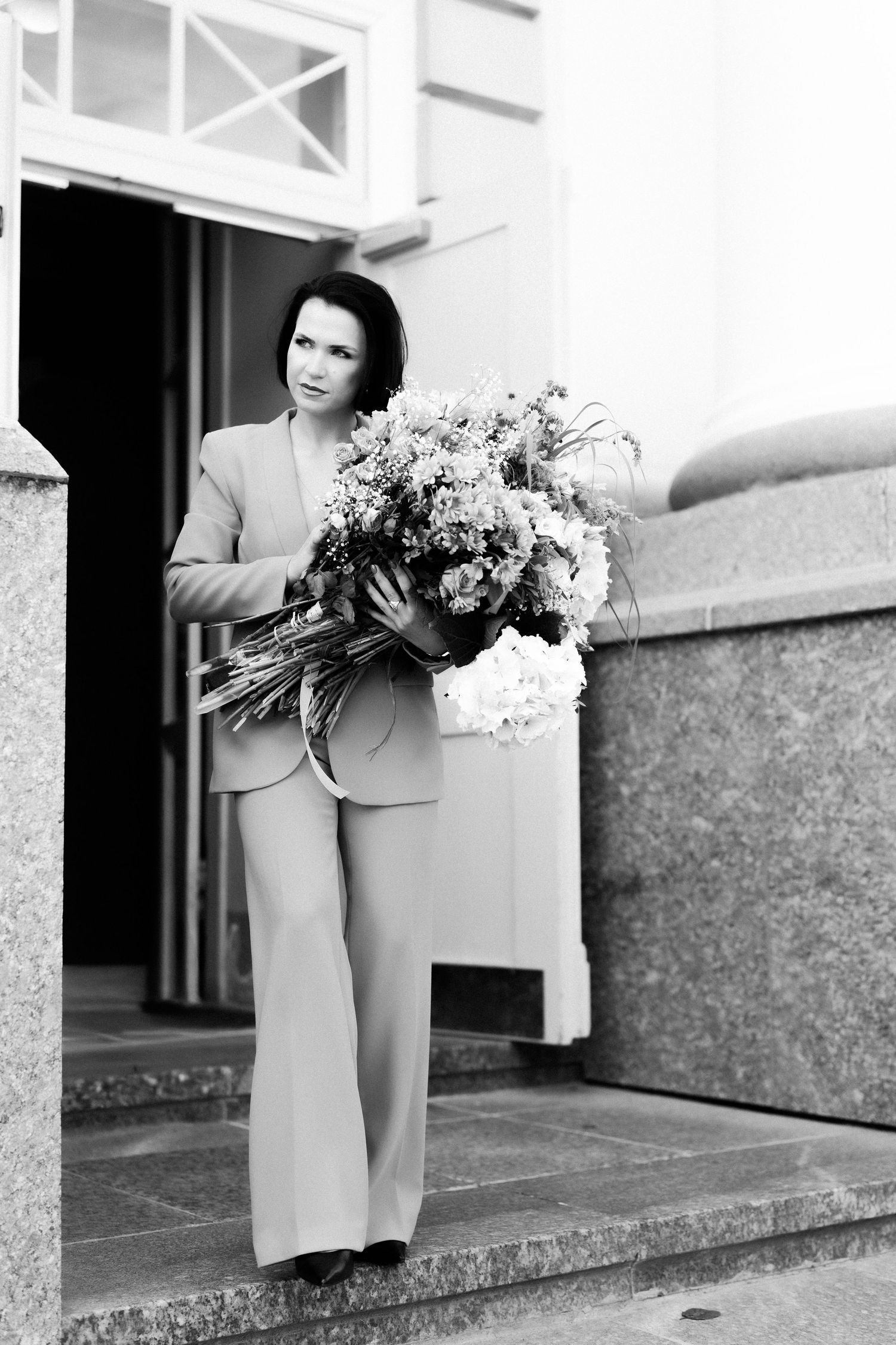 Woman in suit holding large floral bouquet on building steps, black and white photography.