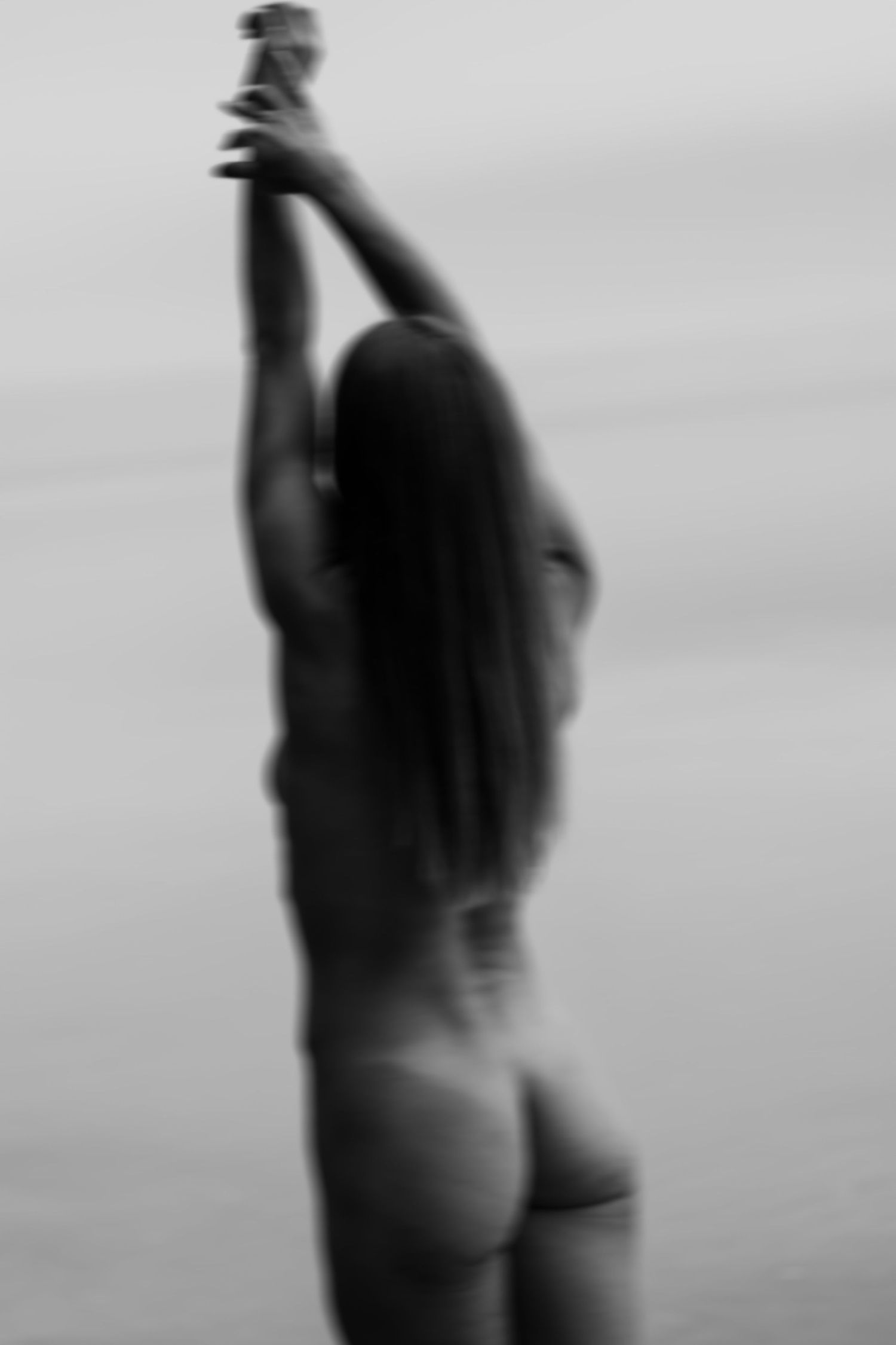 Artistic black and white photograph of a nude silhouette stretching upward against a soft, blurred background.