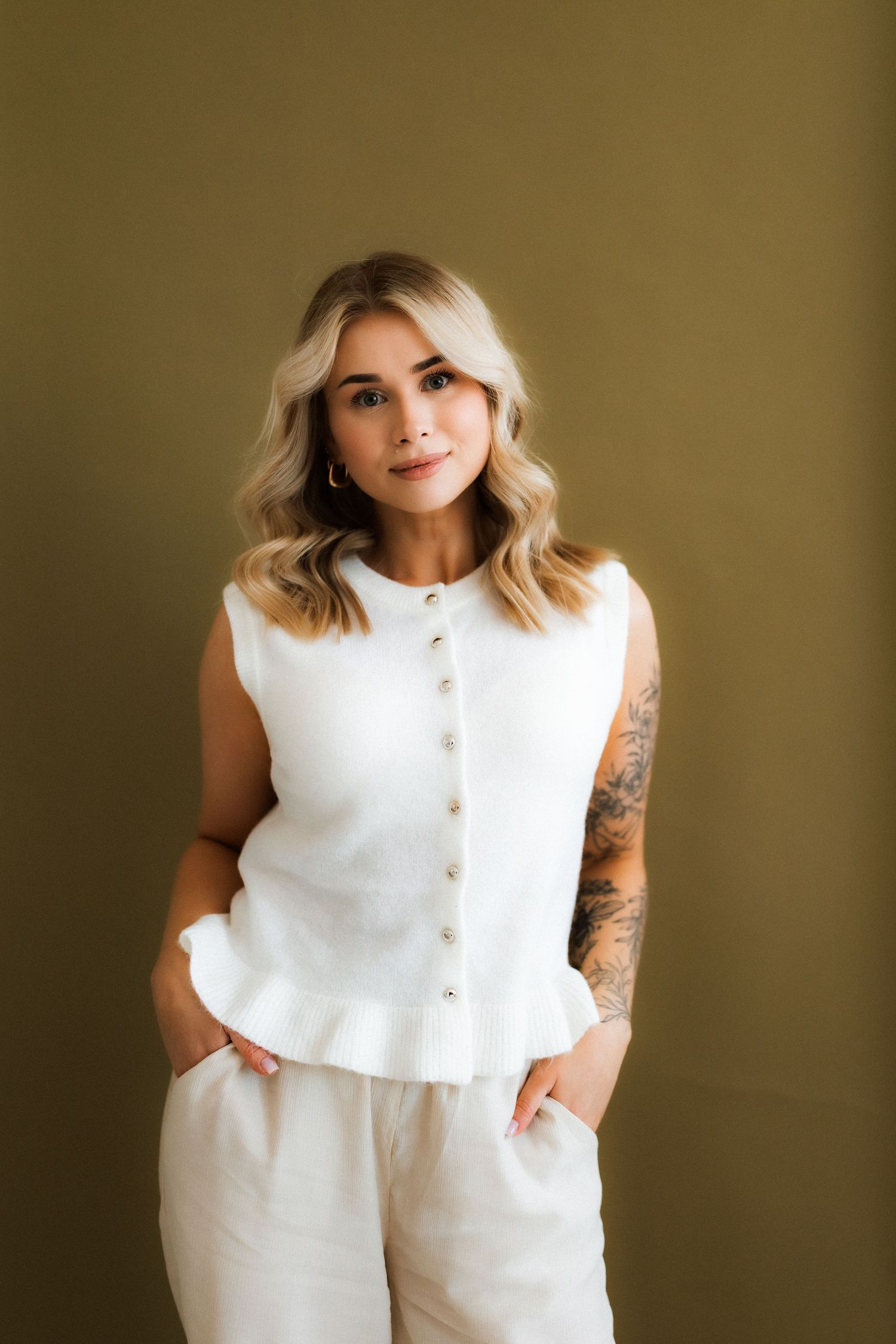 A person in a white sleeveless button-up top and matching pants poses against a brown backdrop with visible arm tattoos.