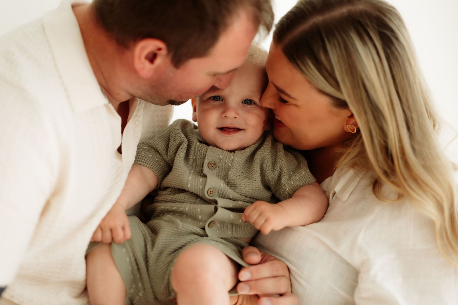 Happy baby in green romper being kissed by smiling parents in bright white studio family portrait photo.