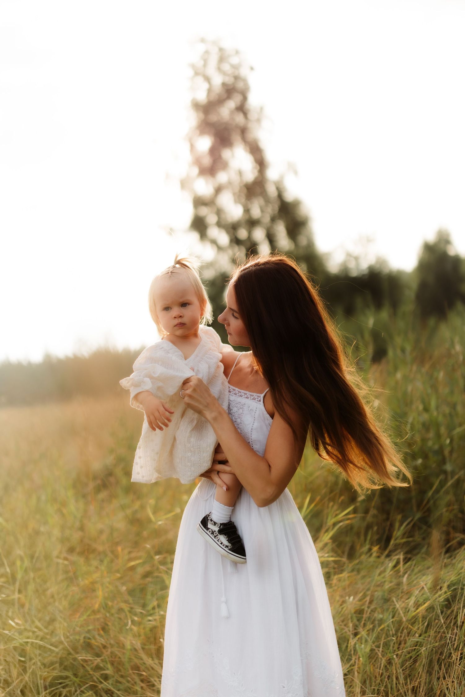 A mother in a white dress holds her baby girl in a sunlit field at golden hour.