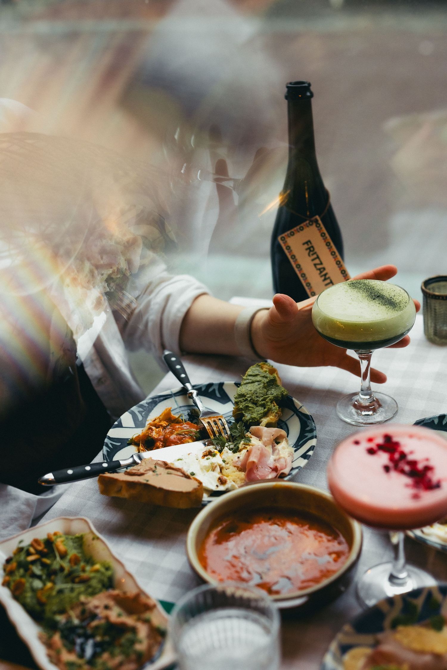 A hand holds a green cocktail over a table filled with colorful dishes, soup, and a wine bottle at a restaurant.