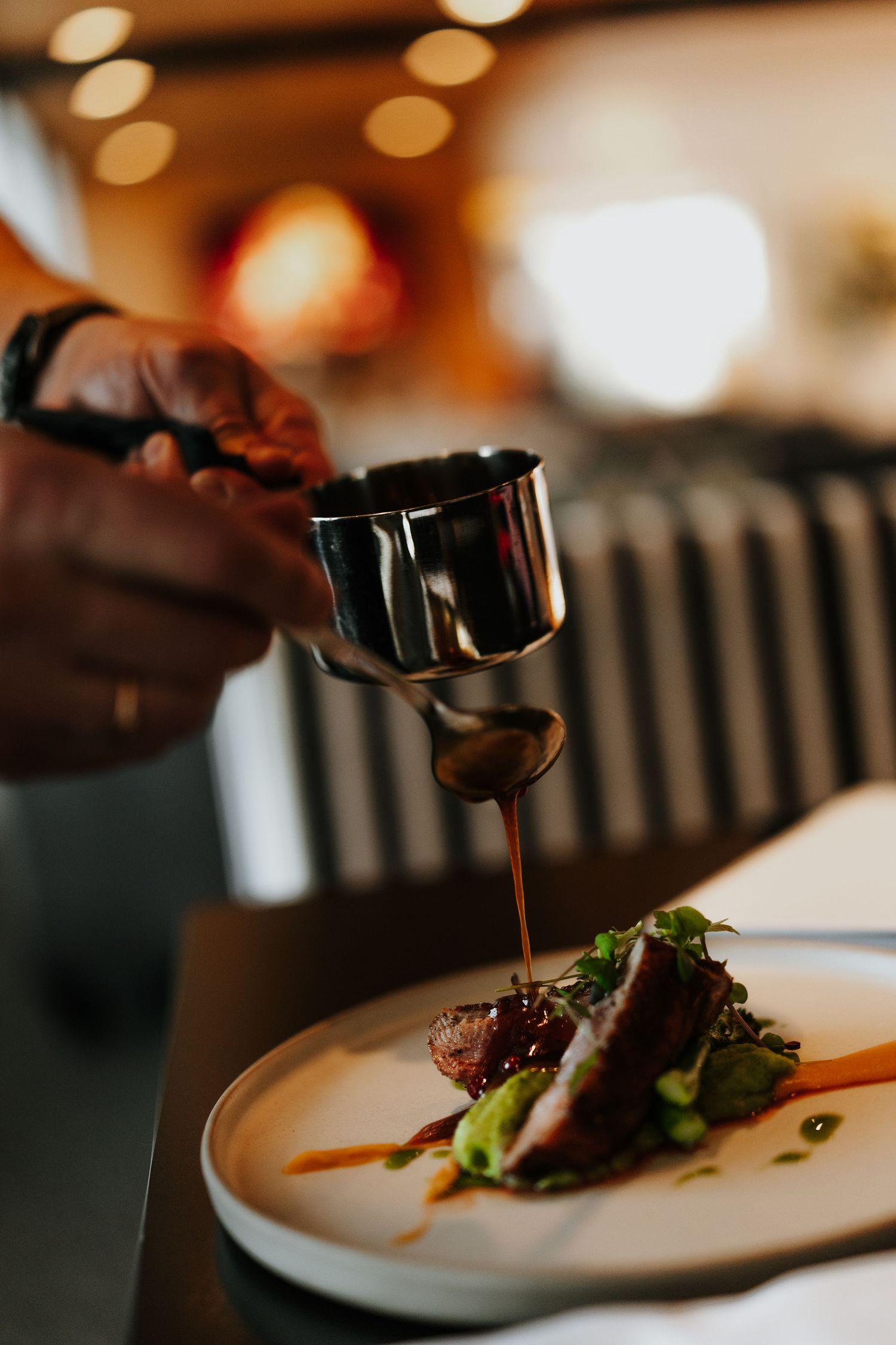 A chef drizzles sauce from a metal container onto a plated gourmet dish in warm restaurant lighting.