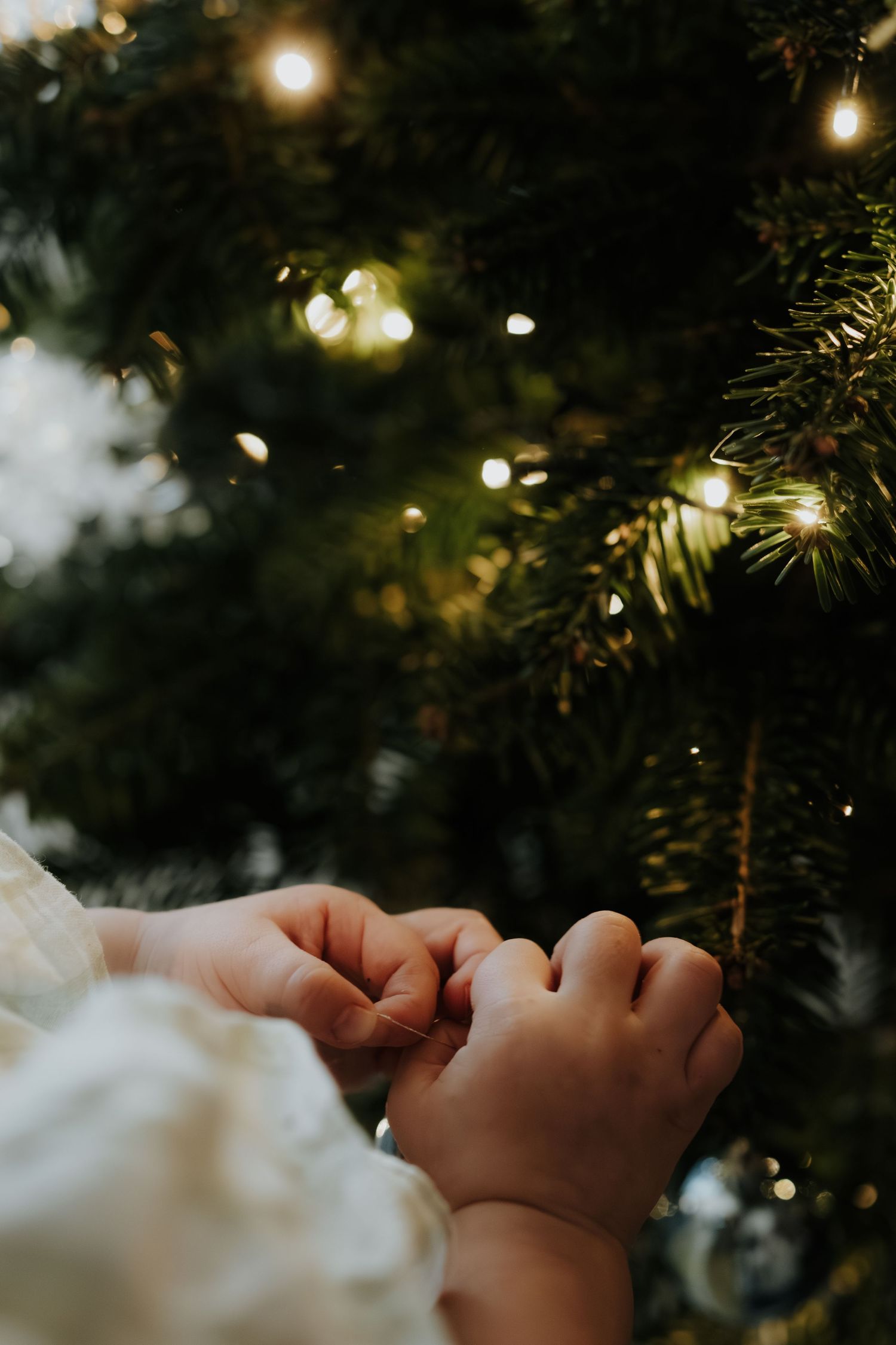 Hand reaching up to touch glowing Christmas tree lights with bokeh effect in dark background.
