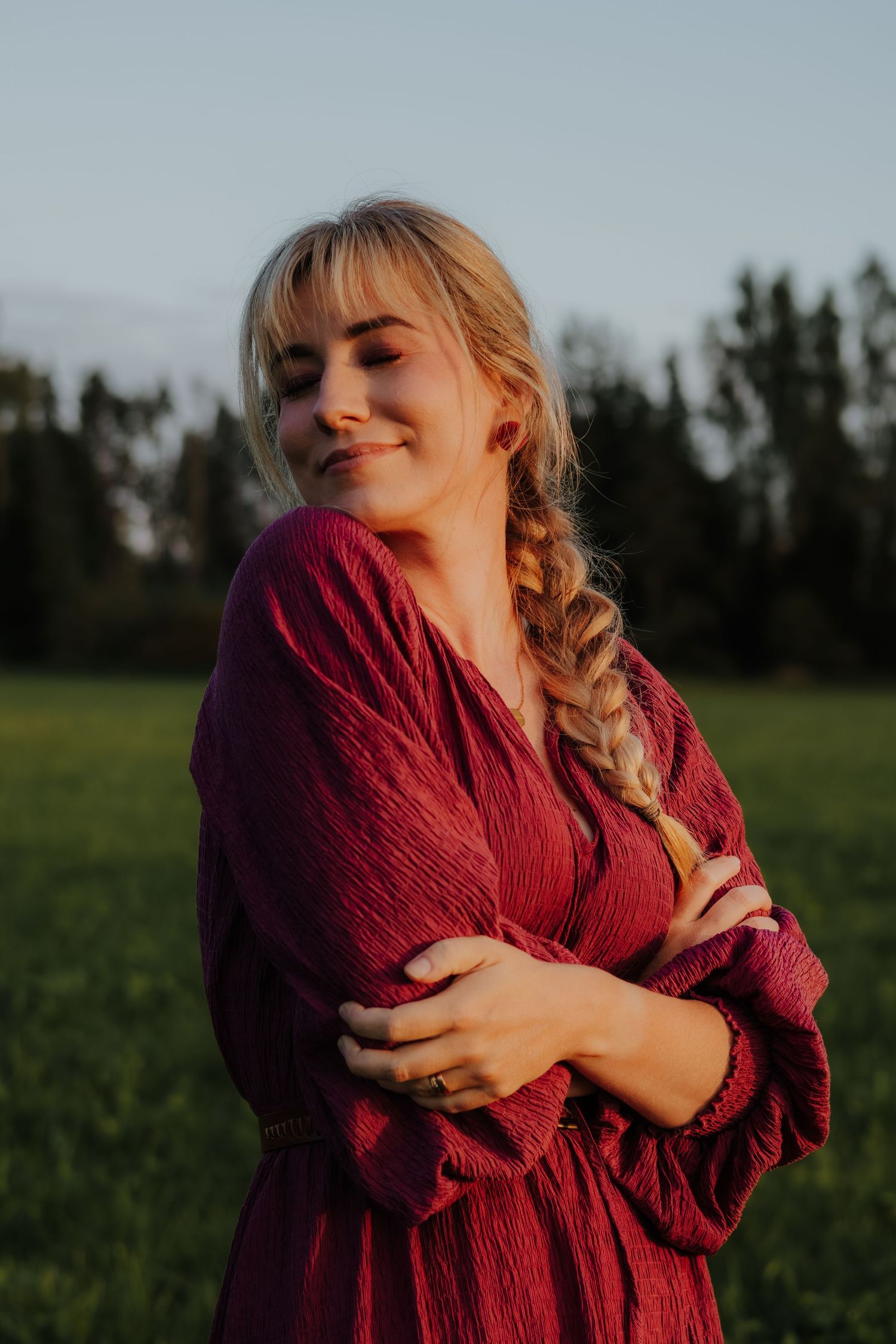 A person wearing a cozy burgundy sweater embraces themselves while standing in a green field at golden hour.