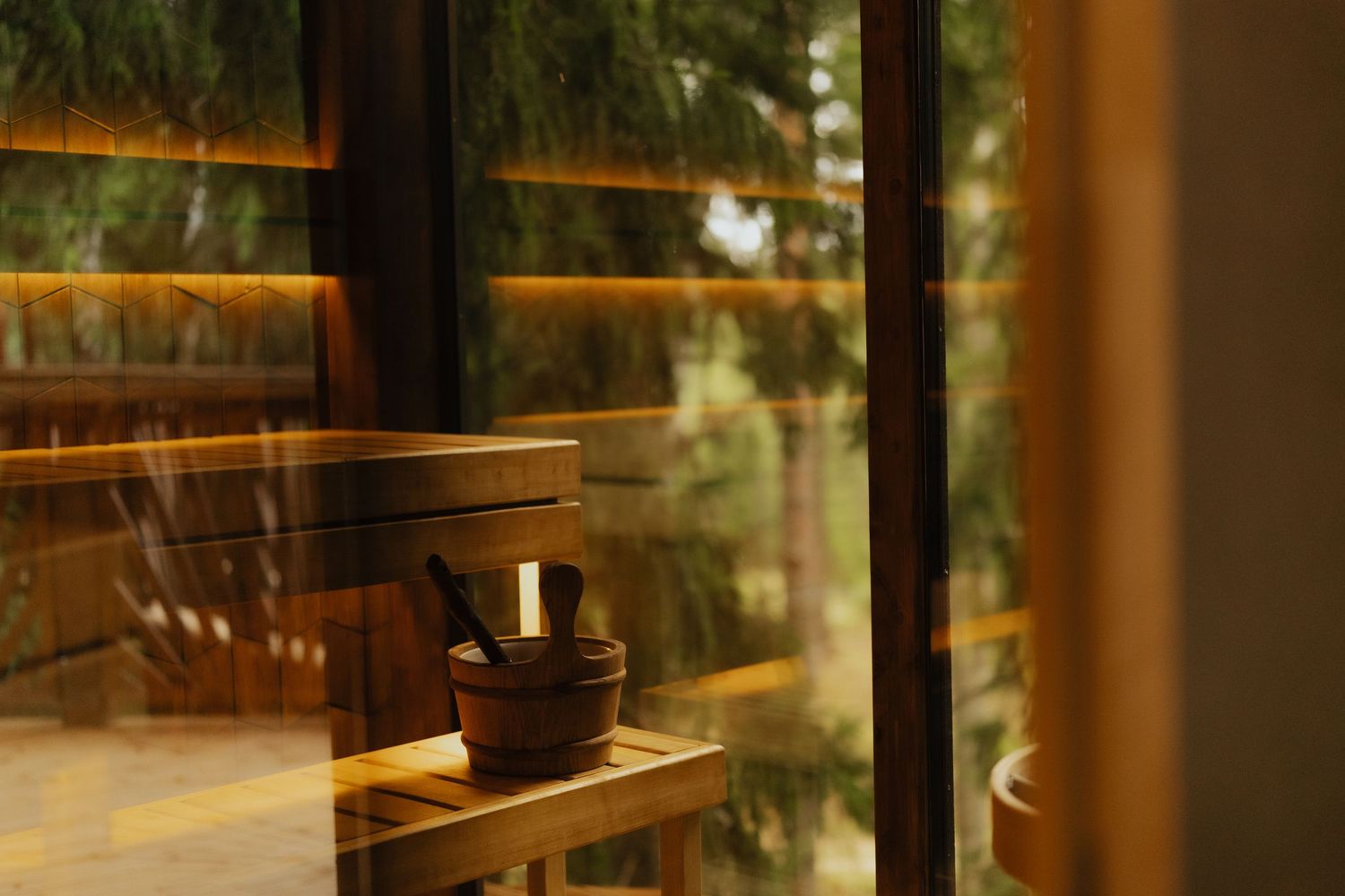 Wooden sauna interior with a small bucket and ladle on a bench beside a large window overlooking lush green trees.