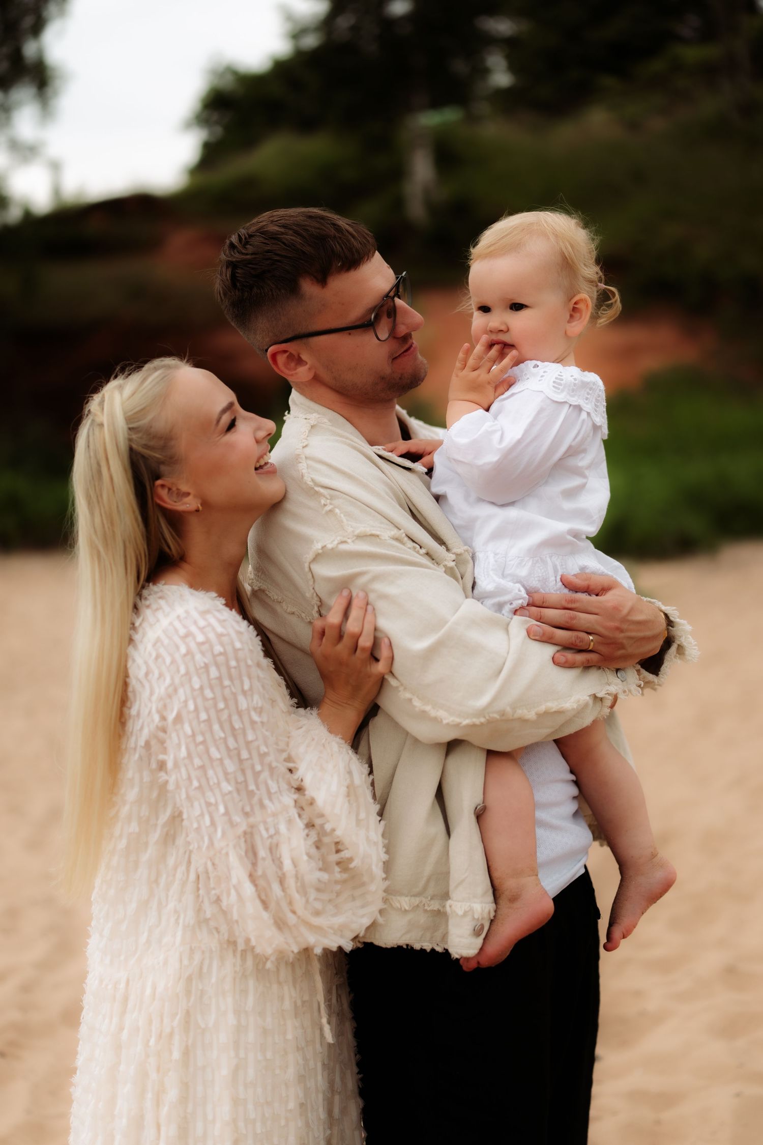 A happy young family with a blonde mother, father in glasses, and baby in white on a sandy beach.