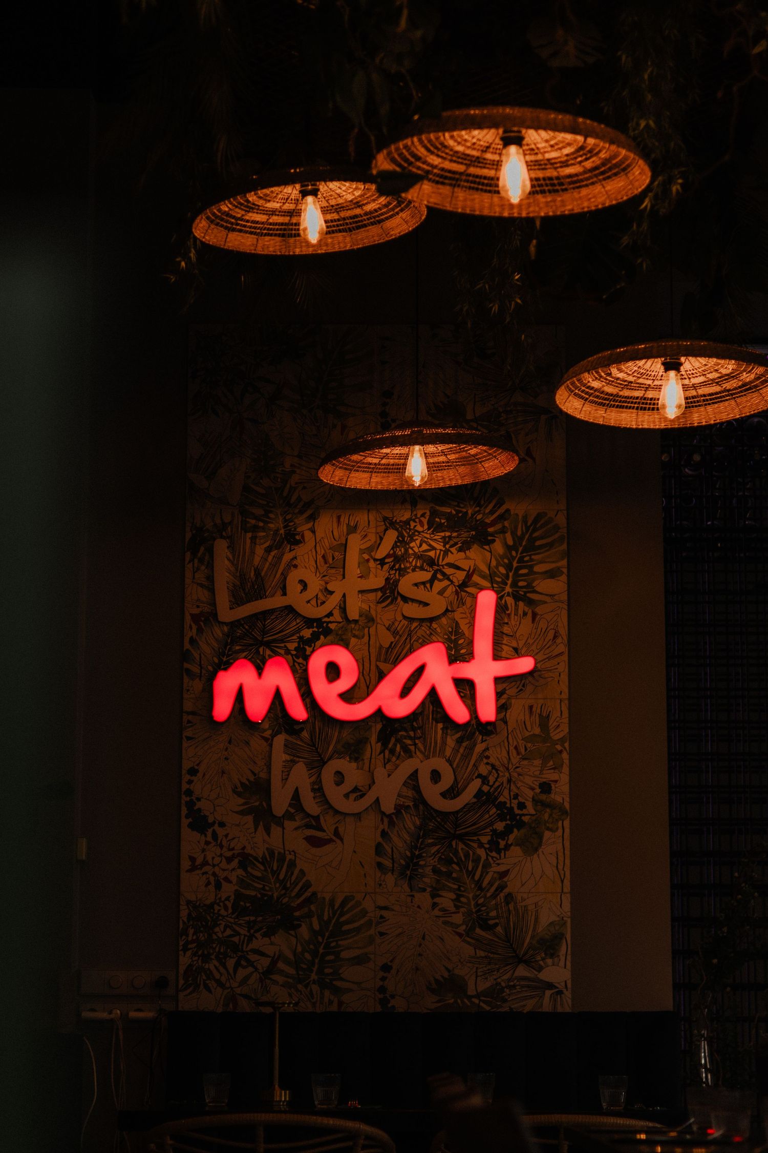 A neon 'meat' sign glows in red against a dark wall with pendant lights hanging above.
