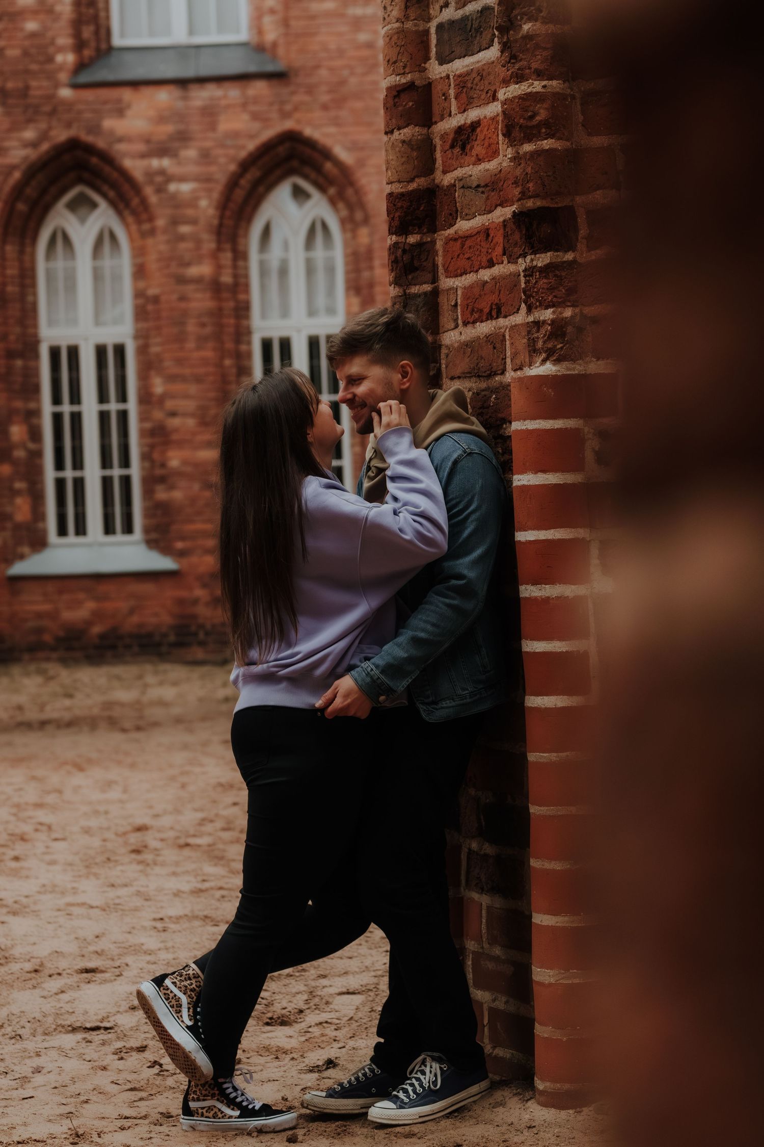Couple in casual wear leaning against red brick wall during romantic outdoor photoshoot.